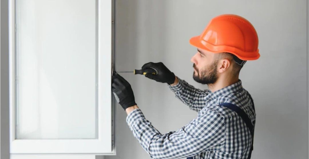 Worker in orange hard hat and gloves using a screwdriver on a window frame.