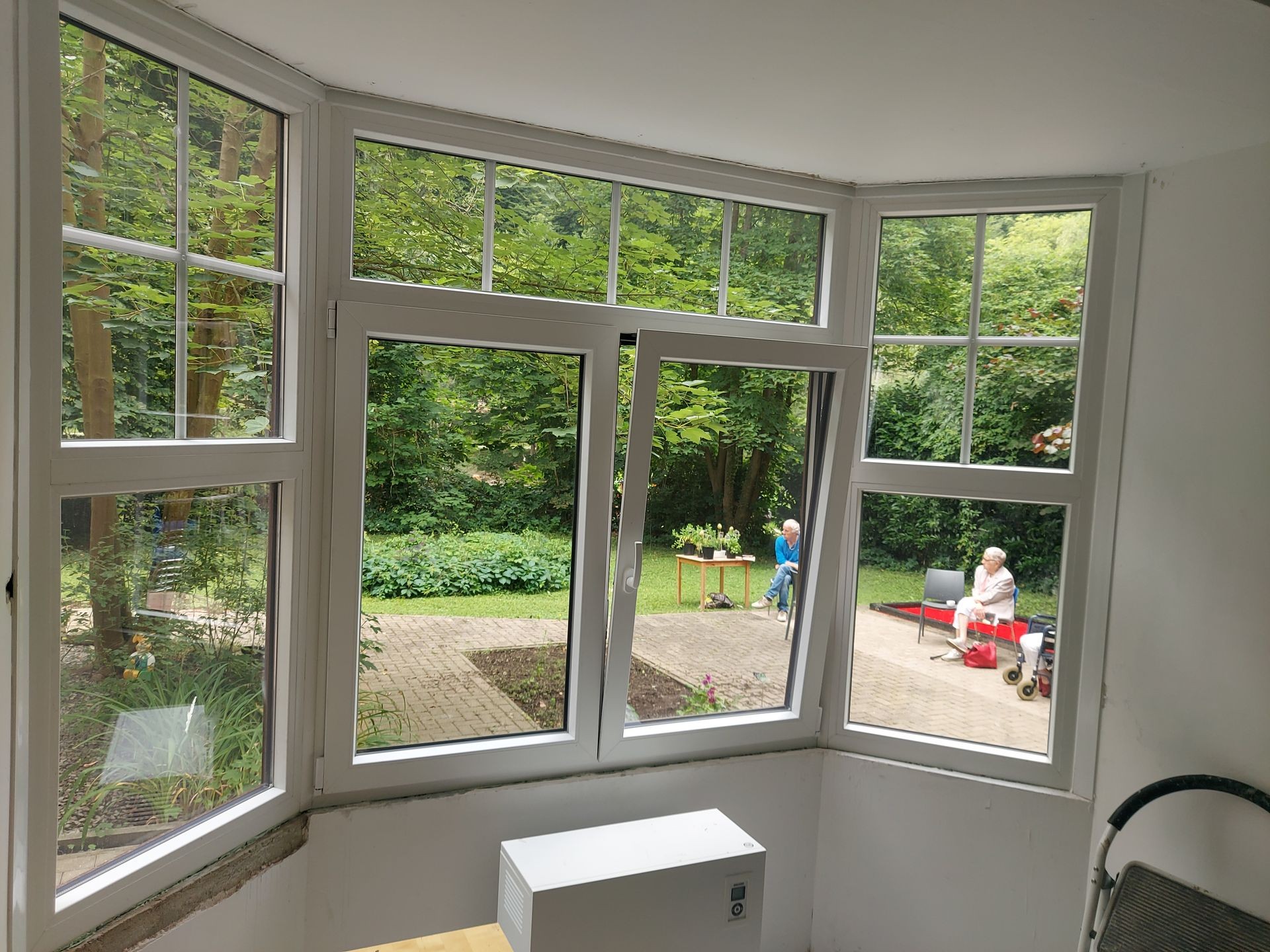 View through bay window of a lush garden with two people sitting outside on a patio.